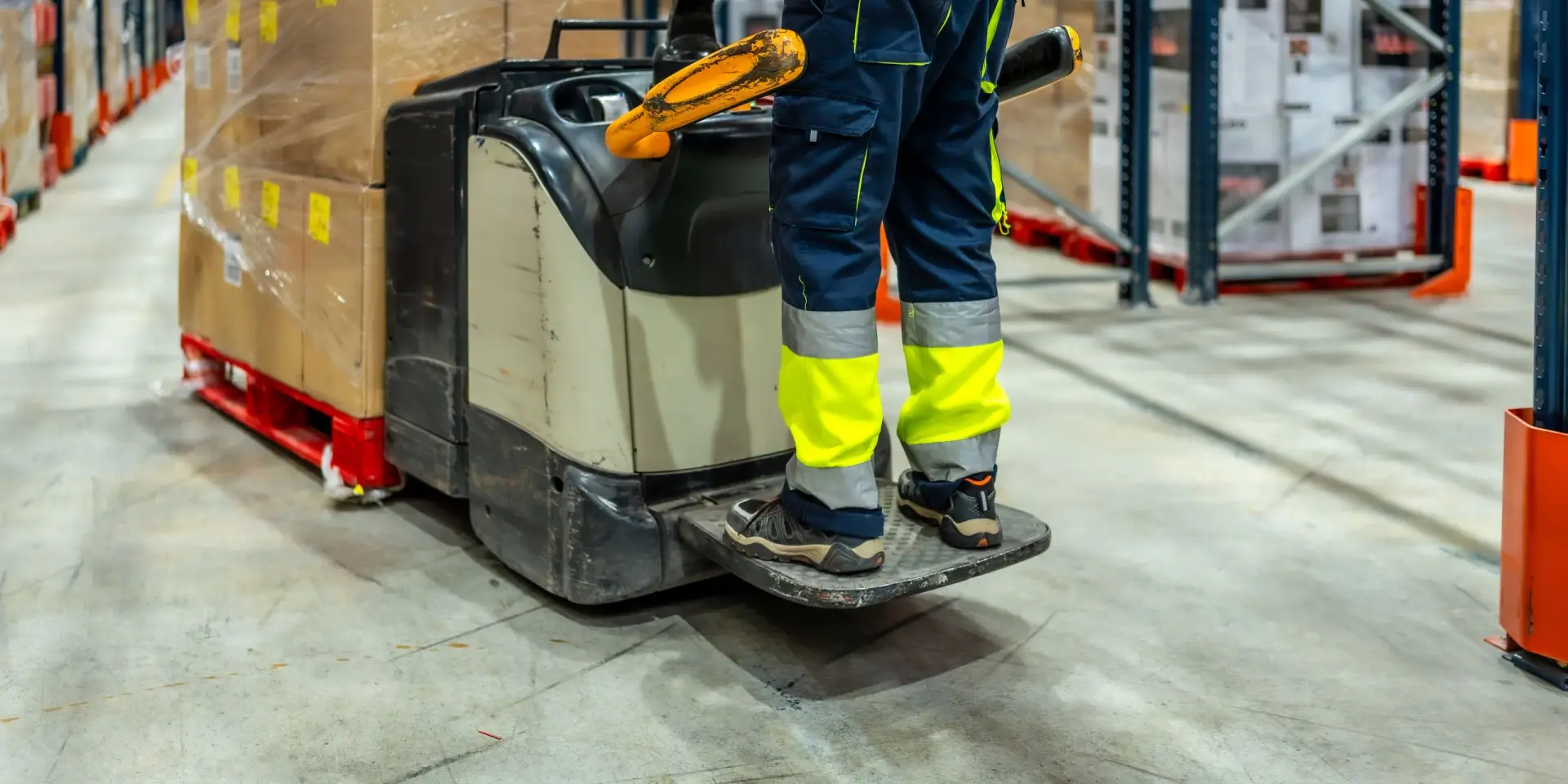 Lagermitarbeiter in Warnkleidung und Sicherheitsschuhen auf der Plattform eines Elektro-Hubwagens zwischen Regalen